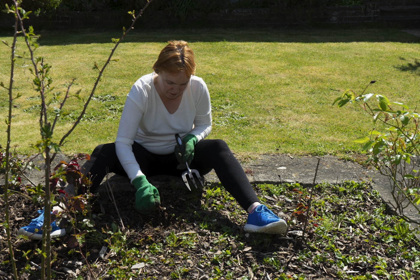 A person wearing green gardening gloves tends a planted bed with a hand fork beside a lawn.